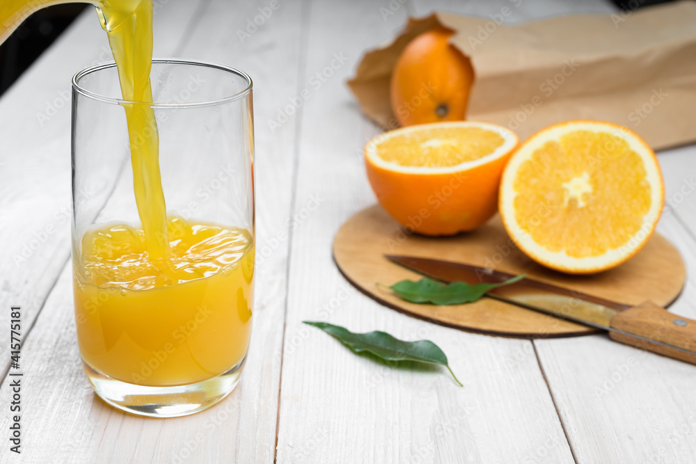 Fototapeta premium Orange juice is poured into a glass on a wooden table, fresh cut oranges in the background