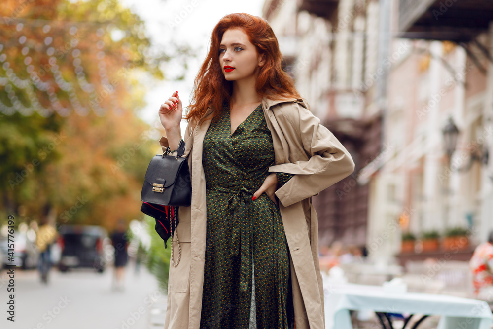Graceful ginger girl in elegant winter outfit walking while holidays in Europe. Stylish leather bag. Beige coat.