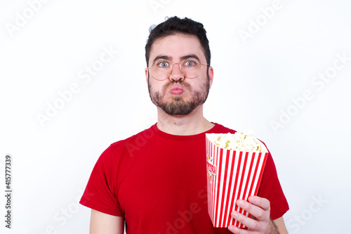 young handsome man in red T-shirt against white wall  puffing cheeks with funny face. Mouth inflated with air, crazy expression.