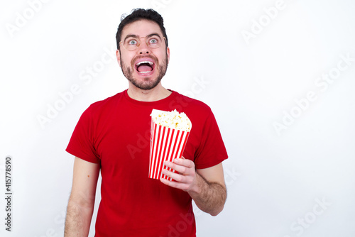Surprised young handsome man in red T-shirt against white background eating popcorn, shrugs shoulders, looking sideways, being happy and excited. Sudden reactions concept.
