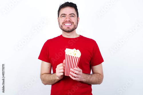 Happy young handsome man in red T-shirt against white background eating popcorn stands against orange studio wall keeps hands on heart, swears be loyal, expresses gratitude. Honesty concept.