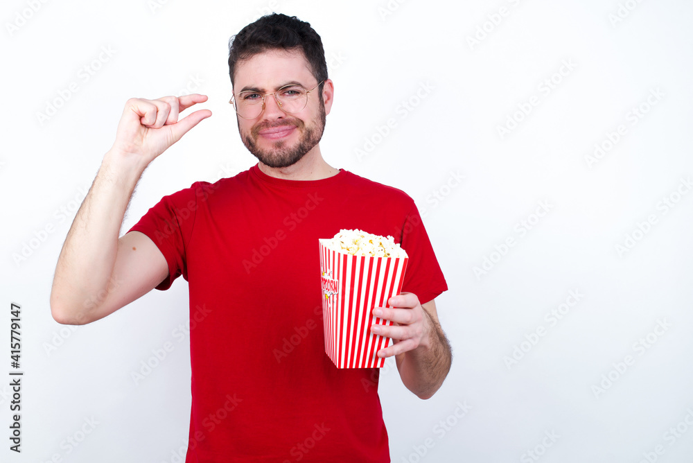 Upset young handsome man in red T-shirt against white background eating popcorn shapes little gesture with hand demonstrates something very tiny small size. Not very much Upset young handsome man in red T-shirt against white background eating popcorn shapes little gesture with hand demonstrates something very tiny small size. Not very much