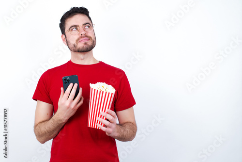 Thinking dreaming Young handsome man in red T-shirt against white background eating popcorn using mobile phone and holding hand on face. Taking decisions and social media concept.