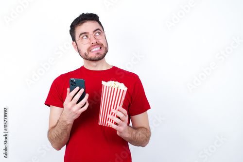 Image of a thinking dreaming Young handsome man in red T-shirt against white background eating popcorn using mobile phone and holding hand on face. Taking decisions and social media concept.
