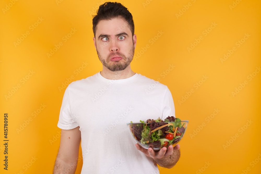 young handsome Caucasian man holding a salad bowl against yellow wall crosses eyes, puts lips, makes grimace with awkward expression has fun alone, plays fool. young handsome Caucasian man holding a salad bowl against yellow wall crosses eyes, puts lips, makes grimace with awkward expression has fun alone, plays fool.