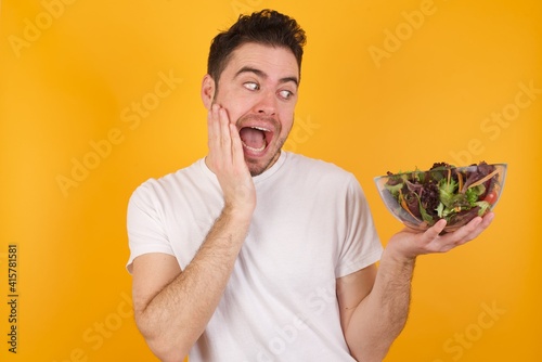 young handsome Caucasian man holding a salad bowl against yellow wall excited looking to the side hand on face. Advertisement and amazement concept.