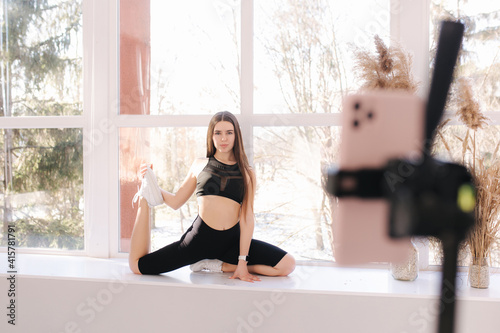 Young athletic woman blogger in balck sportswear shoots video on phone as she does exercises at home in living room on windowsill. Sport and recreation concept. Fitness vlogger recording live tutorial