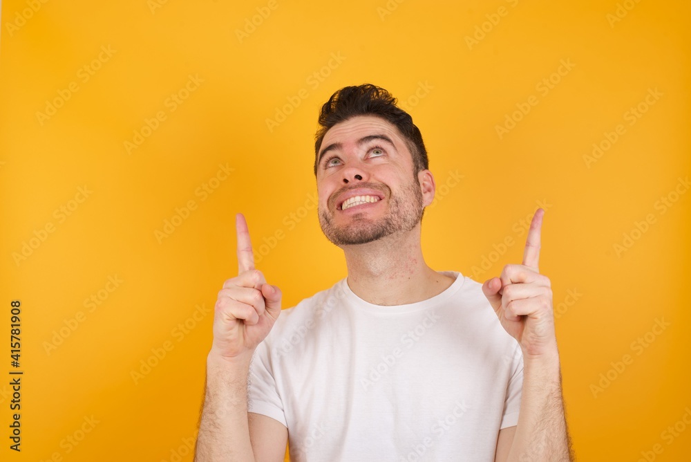 Successful friendly looking young handsome Caucasian man against yellow wall exclaiming excitedly, pointing both index fingers up, indicating something. Successful friendly looking young handsome Caucasian man against yellow wall exclaiming excitedly, pointing both index fingers up, indicating something.