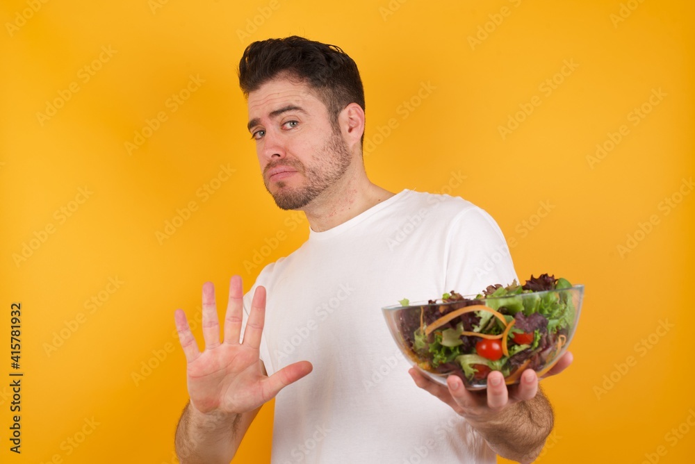 Afraid young handsome Caucasian man holding a salad bowl against yellow wall, makes terrified expression and stop gesture with both hands saying: Stay there. Panic concept.
