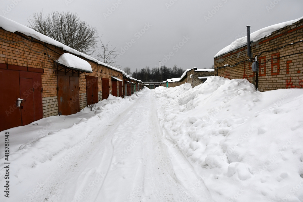 A cloudy winter day. Snowfall. Rows of one-story brick garages with closed metal painted gates. Drifts of snow near the walls along the road. Snow on the roofs.