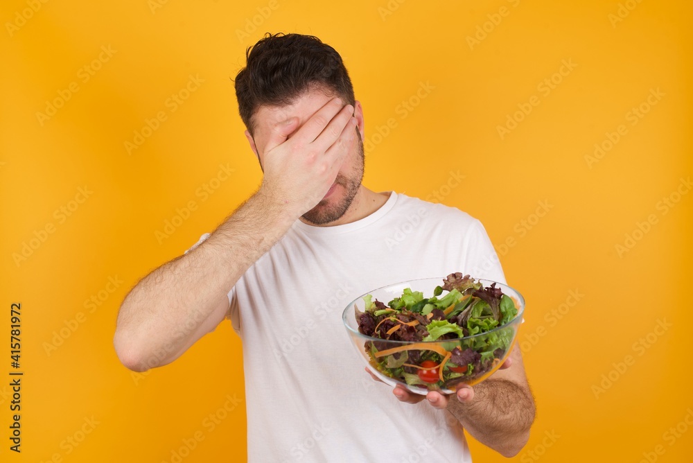young handsome Caucasian man holding a salad bowl against yellow wall making facepalm gesture while smiling amazed with stupid situation.
