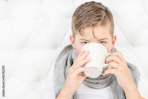 A sick child lies on a bed in a warm scarf and drinks hot tea from a mug in the beadroom. An unhealthy child drinks from mug. Cold season.