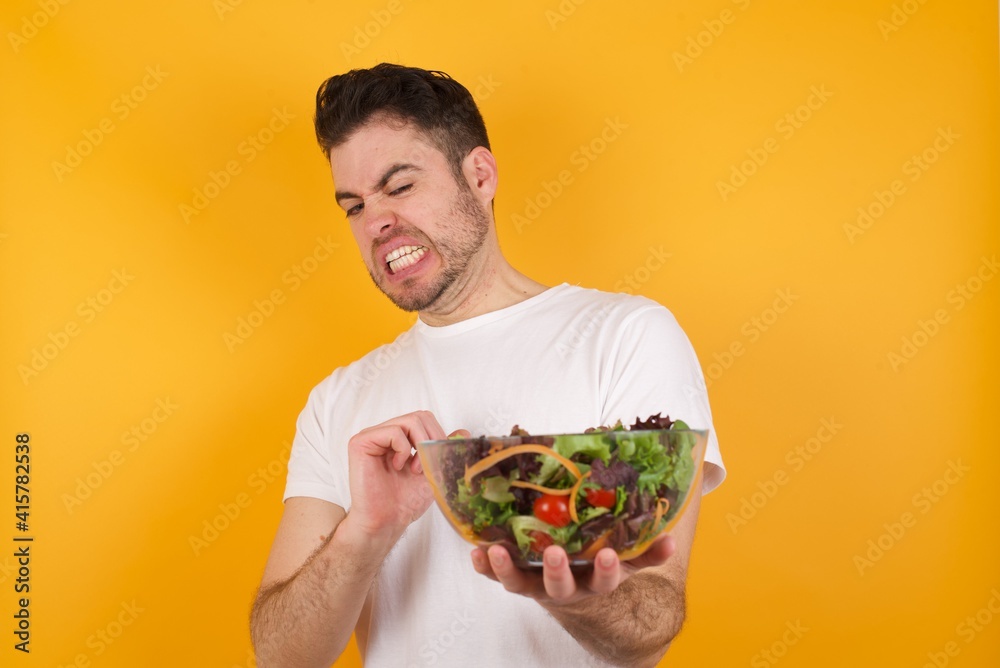 Ugh how disgusting! Displeased young handsome Caucasian man holding a salad bowl against yellow wall, has dissatisfied facial expression as sees something abominable. Ugh how disgusting! Displeased young handsome Caucasian man holding a salad bowl against yellow wall, has dissatisfied facial expression as sees something abominable.