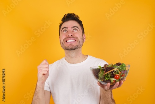 young handsome Caucasian man holding a salad bowl against yellow wall having success being glad to achieve his goals. Victory and triumph concept.