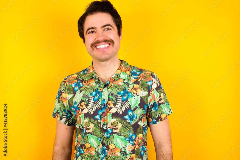 Portrait of successful young handsome Caucasian man wearing Hawaiian shirt against yellow wall, smiling broadly with self-assured expression. Confidence and business concept. Portrait of successful young handsome Caucasian man wearing Hawaiian shirt against yellow wall, smiling broadly with self-assured expression. Confidence and business concept.