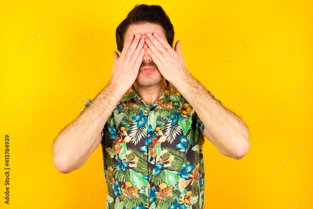 young handsome Caucasian man wearing Hawaiian shirt against yellow wall covering eyes with both hands, doesn't want to see anything or feeling ashamed. Human feelings reactions.