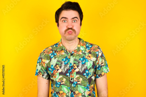 Wallpaper Mural young handsome Caucasian man wearing Hawaiian shirt against yellow wall puffing cheeks with funny face. Mouth inflated with air, crazy expression. Torontodigital.ca
