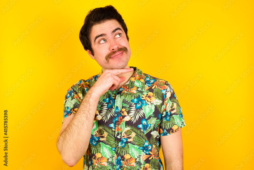 custom made wallpaper toronto digitalDreamy young handsome Caucasian man wearing Hawaiian shirt against yellow wall with pleasant expression, looks sideways, keeps hand under chin, thinks about something pleasant.