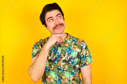 Wallpaper Mural Dreamy young handsome Caucasian man wearing Hawaiian shirt against yellow wall with pleasant expression, looks sideways, keeps hand under chin, thinks about something pleasant. Torontodigital.ca