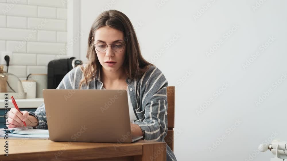 A serious young woman wearing glasses is writing something while working with her laptop computer sitting at the table in the kitchen at home A serious young woman wearing glasses is writing something while working with her laptop computer sitting at the table in the kitchen at home