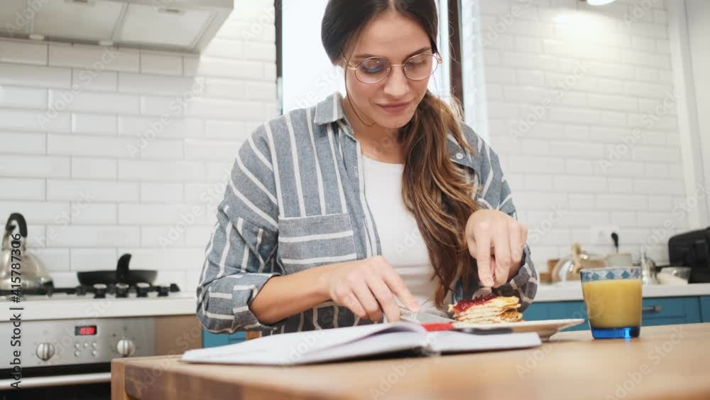 A pleased young woman wearing glasses is eating breakfast in the kitchen at home A pleased young woman wearing glasses is eating breakfast in the kitchen at home