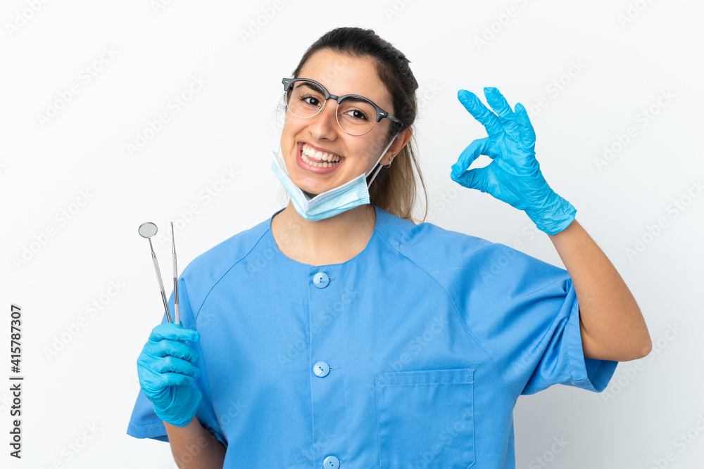 Young woman dentist holding tools isolated on white background showing ok sign with fingers Young woman dentist holding tools isolated on white background showing ok sign with fingers