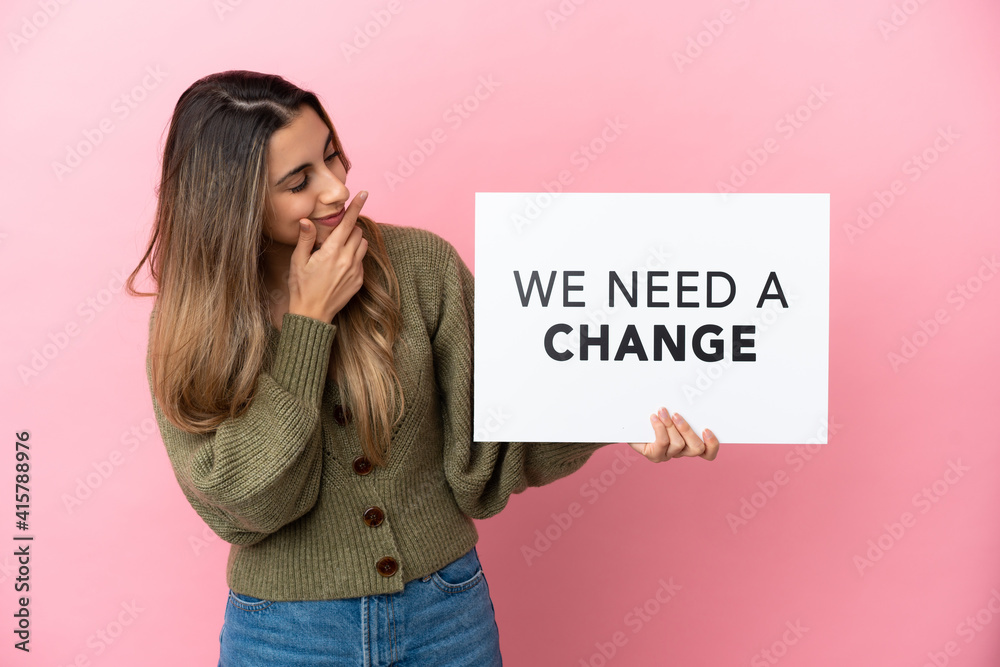 Young caucasian woman isolated on pink background holding a placard with text We Need a Change and thinking Young caucasian woman isolated on pink background holding a placard with text We Need a Change and thinking