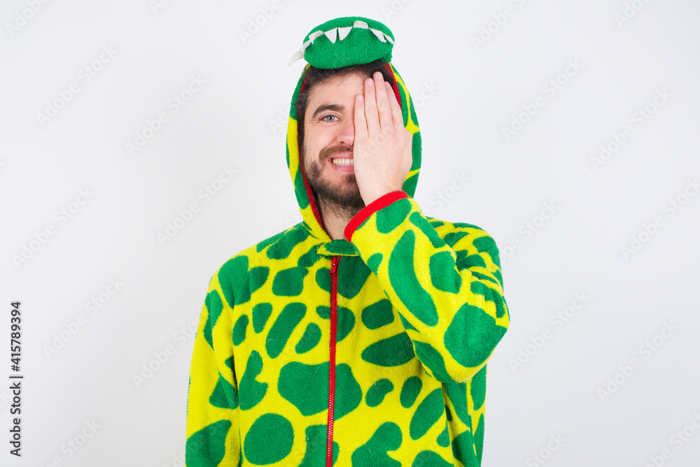 Fototapeta premium young caucasian man wearing a pajama standing against white wall covering one eye with her hand, confident smile on face and surprise emotion.