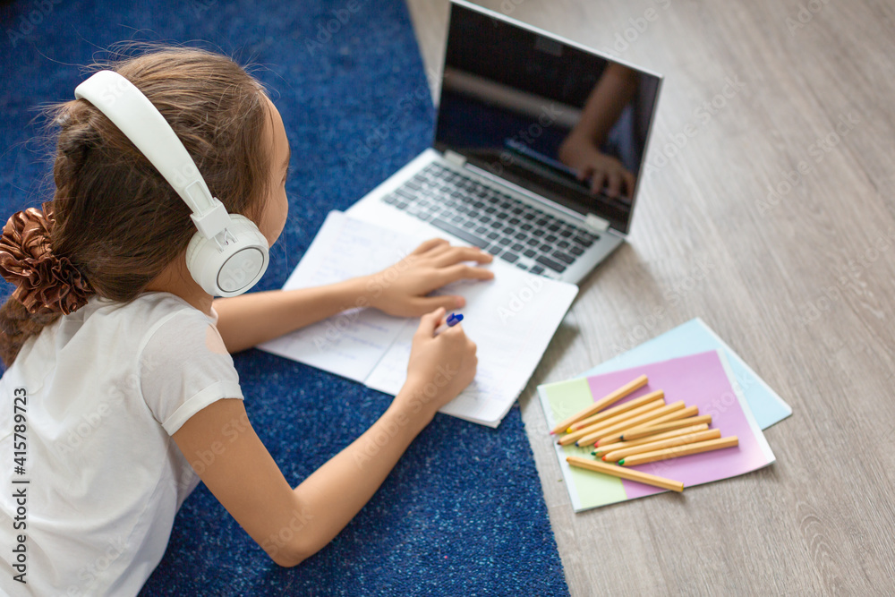 Fototapeta premium Pupil girl doing her homework online in front of a laptop monitor lying on the floor at home.