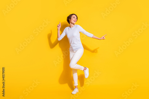 Full length photo of cute funny young woman wear white shirt dancing pointing empty space isolated yellow color background