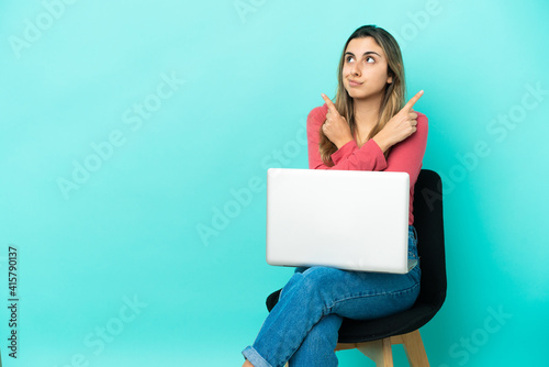 Young caucasian woman sitting on a chair with her pc isolated on blue background pointing to the laterals having doubts © luismolinero