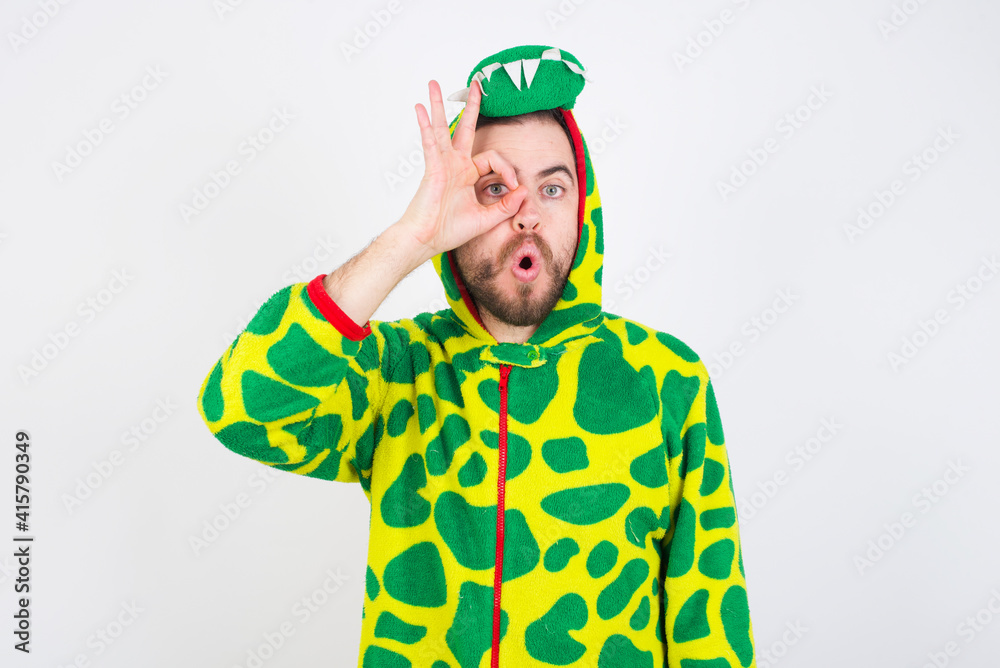 young caucasian man wearing a pajama standing against white wall doing ok gesture shocked with surprised face, eye looking through fingers. Unbelieving expression. young caucasian man wearing a pajama standing against white wall doing ok gesture shocked with surprised face, eye looking through fingers. Unbelieving expression.