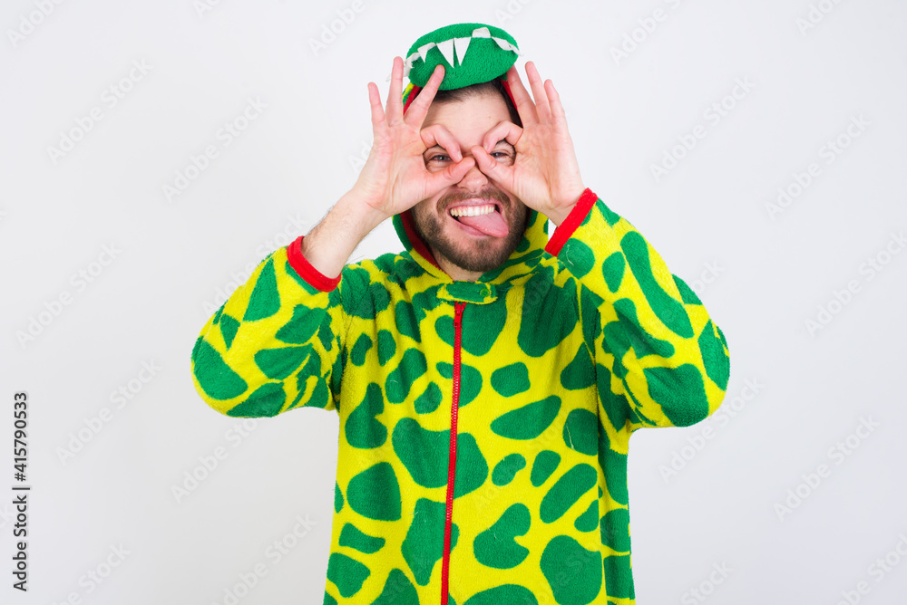 young caucasian man wearing a pajama standing against white wall doing ok gesture like binoculars sticking tongue out, eyes looking through fingers. Crazy expression. young caucasian man wearing a pajama standing against white wall doing ok gesture like binoculars sticking tongue out, eyes looking through fingers. Crazy expression.