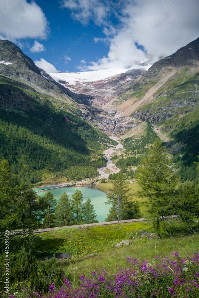Fototapeta premium Scenic view of mountains against sky. Majestic waterfall leading from melting snow near Alp Grum. Rhaetian Railway track in foreground.