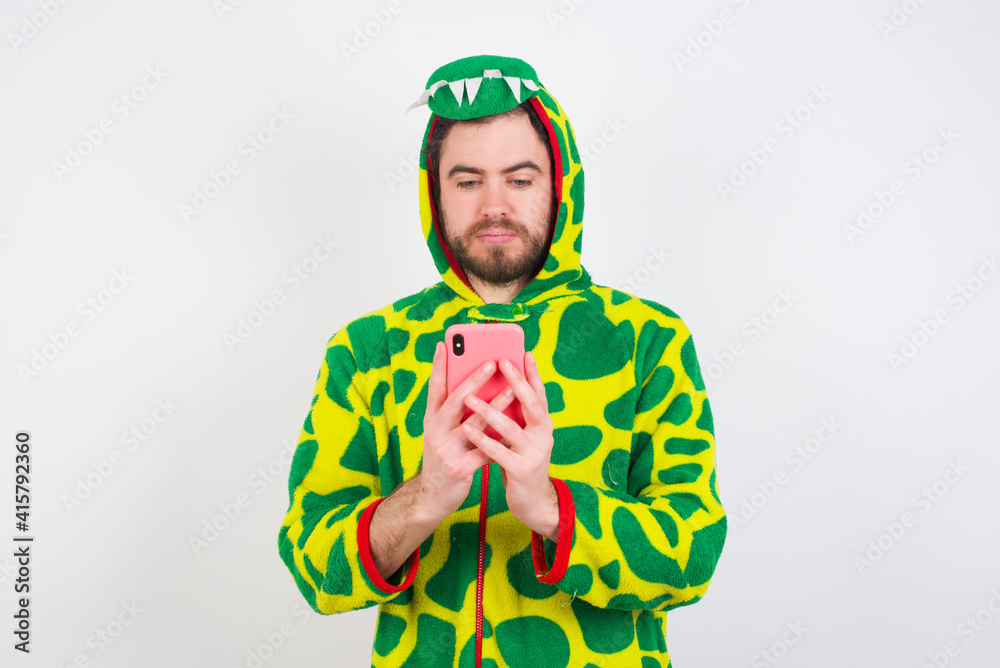 Fototapeta premium Excited young caucasian man wearing a pajama standing against white background winking and eye hold smart phone use read social network news