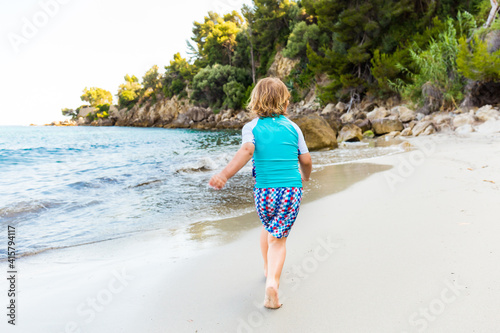 Child playing on beautiful summer beach. Little blond boy wearing rash guard swimsuit running at sea shore. Ocean vacation with kid. Water fun. Family holiday on tropical island. © Anna