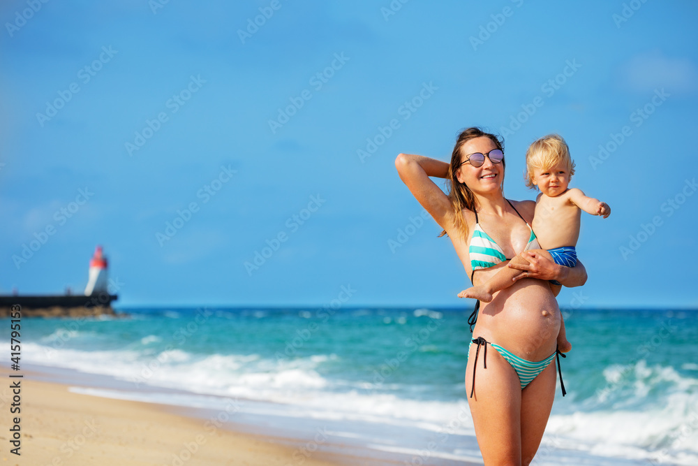 Happy laughing portrait of a mother hold little blond smiling toddler pointing with finger boy on the beach near lighthouse