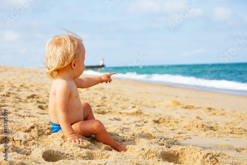 Portrait of a little calm blond toddler boy sitting and point to the sea on the beach near lighthouse during summer vacations