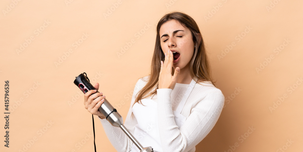Obraz premium Woman using hand blender over isolated background yawning and covering wide open mouth with hand