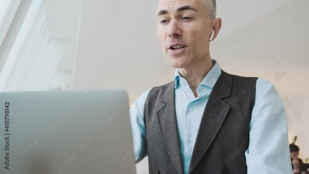 A handsome man wearing earbuds is talking with video-connection using his laptop computer sitting inside the public space A handsome man wearing earbuds is talking with video-connection using his laptop computer sitting inside the public space