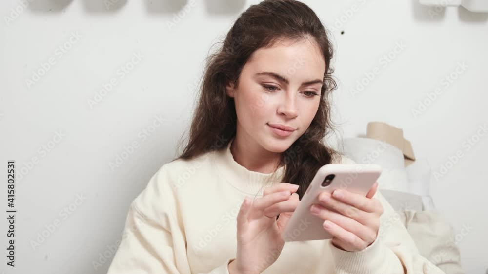 A smiling young woman seamstress is using her smartphone sitting at the design studio A smiling young woman seamstress is using her smartphone sitting at the design studio