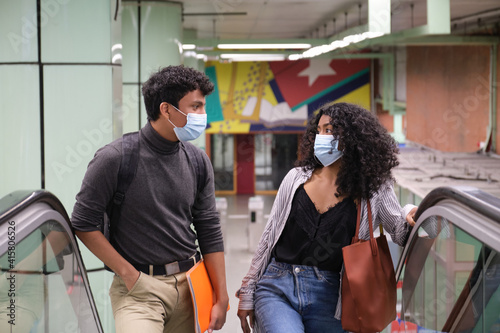 Young latin couple wearing protective face mask walking up into the escalators at the train or metro station. New normal at public transport.