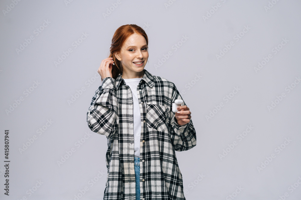 Happy young woman putting wireless earphones on isolated white background, looking at camera. Pretty redhead lady model wearing casual fashion clothes emotionally showing facial expressions, copyspace Happy young woman putting wireless earphones on isolated white background, looking at camera. Pretty redhead lady model wearing casual fashion clothes emotionally showing facial expressions, copyspace
