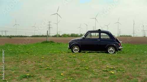 A fiat 500 car parked on a grassy field with windwheels on a cloudy day, tracking shot photo