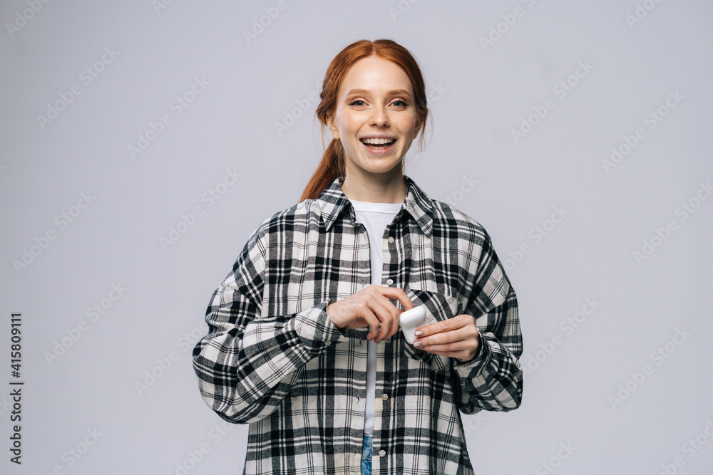 Cheerful young woman taking wireless earphones out of case on isolated white background, looking at camera. Pretty redhead lady model in casual fashion clothes emotionally showing facial expressions. Cheerful young woman taking wireless earphones out of case on isolated white background, looking at camera. Pretty redhead lady model in casual fashion clothes emotionally showing facial expressions.