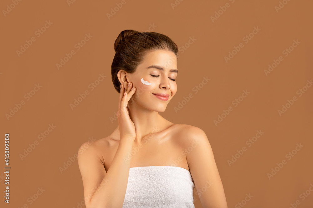 Obraz premium Beauty and wellness concept. Lovely young woman applying face cream over brown studio background