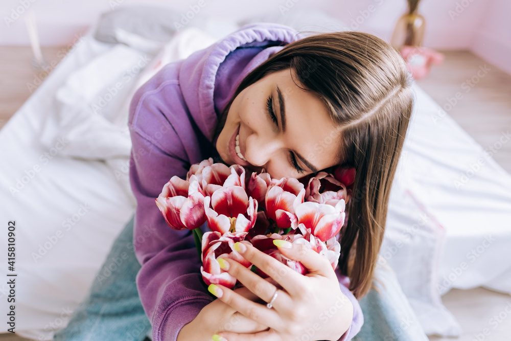 Young caucasian happy woman smelling bouquet of fresh tulips at home. Sitting on the bed in spring morning, woman day concept. Young caucasian happy woman smelling bouquet of fresh tulips at home. Sitting on the bed in spring morning, woman day concept.