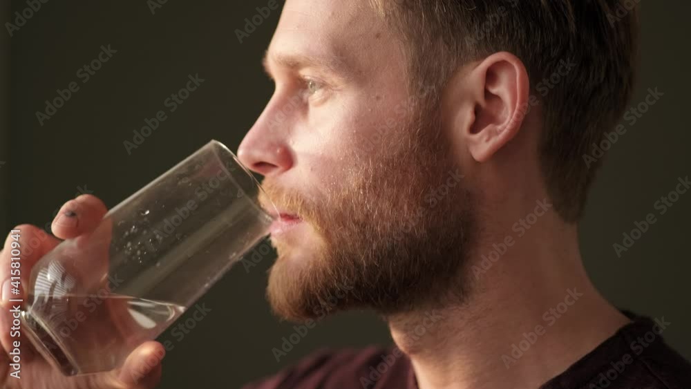 A close-up side view of a pleased man is drinking fresh water while standing in the kitchen at home in the morning A close-up side view of a pleased man is drinking fresh water while standing in the kitchen at home in the morning