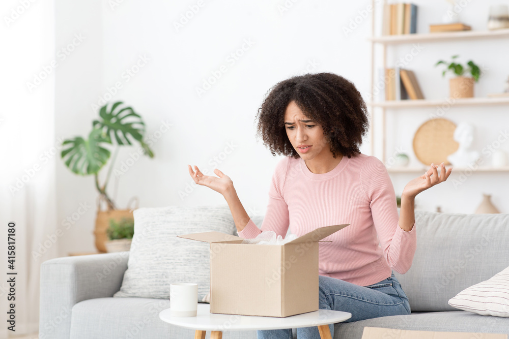 Upset millennial african american woman looks into cardboard box with purchases Upset millennial african american woman looks into cardboard box with purchases