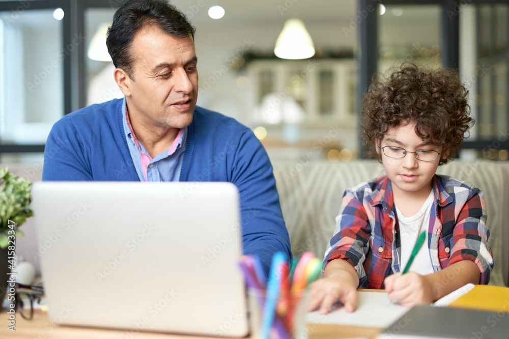 Father can help. Portrait of middle aged hispanic father spending time with his son. Little boy sitting at the desk together with his dad and doing homework Father can help. Portrait of middle aged hispanic father spending time with his son. Little boy sitting at the desk together with his dad and doing homework